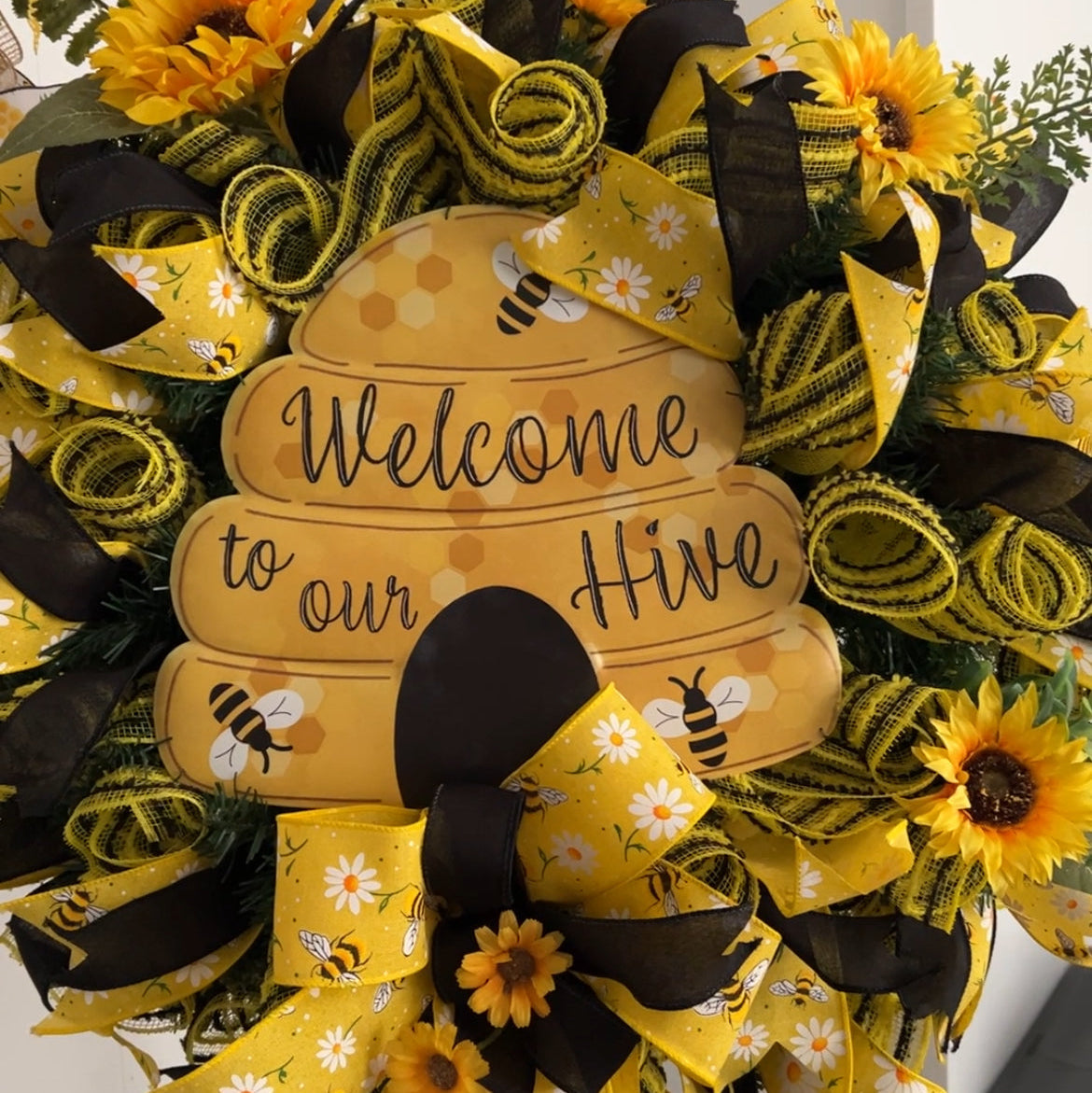 Handmade bee and sunflower wreath featuring a Welcome to Our Hive sign, yellow and black ribbons, daisy accents, and bright sunflowers for summer front door decor.
