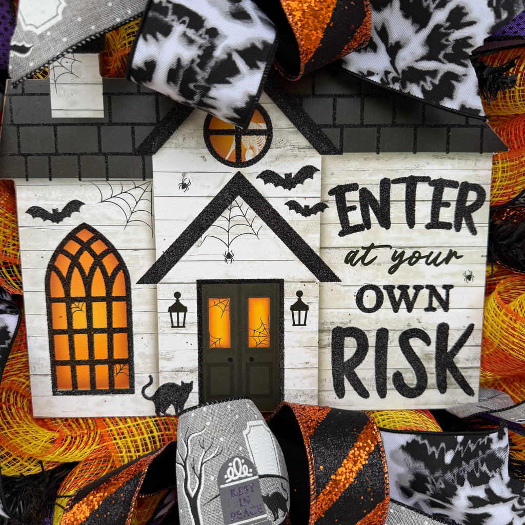 A Halloween-themed decorative sign shaped like a small haunted house, surrounded by layered orange, black, white, and purple mesh ribbon with spiderwebs, bats, tombstones, and the phrase “ENTER at your OWN RISK.”.