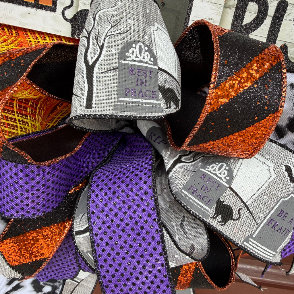 A Halloween-themed decorative wreath or bow made of layered ribbons, featuring gray fabric with black cat and tombstone graphics, black and orange glitter, purple ribbon with black dots, and orange/black striped ribbons.
