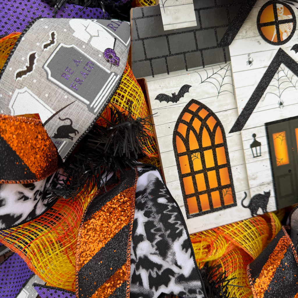 A close-up of Halloween-themed decorative mesh ribbon and a small printed house ornament. The designs feature orange, black, and purple patterns, including stripes, glitter textures, spiderwebs, bats, and a tombstone motif.