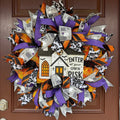 A Halloween-themed mesh wreath hanging on a brown front door. The wreath features purple, orange, black, and white ribbons with a central white house sign that reads 'ENTER at your OWN RISK' with a pumpkin and bats detail.