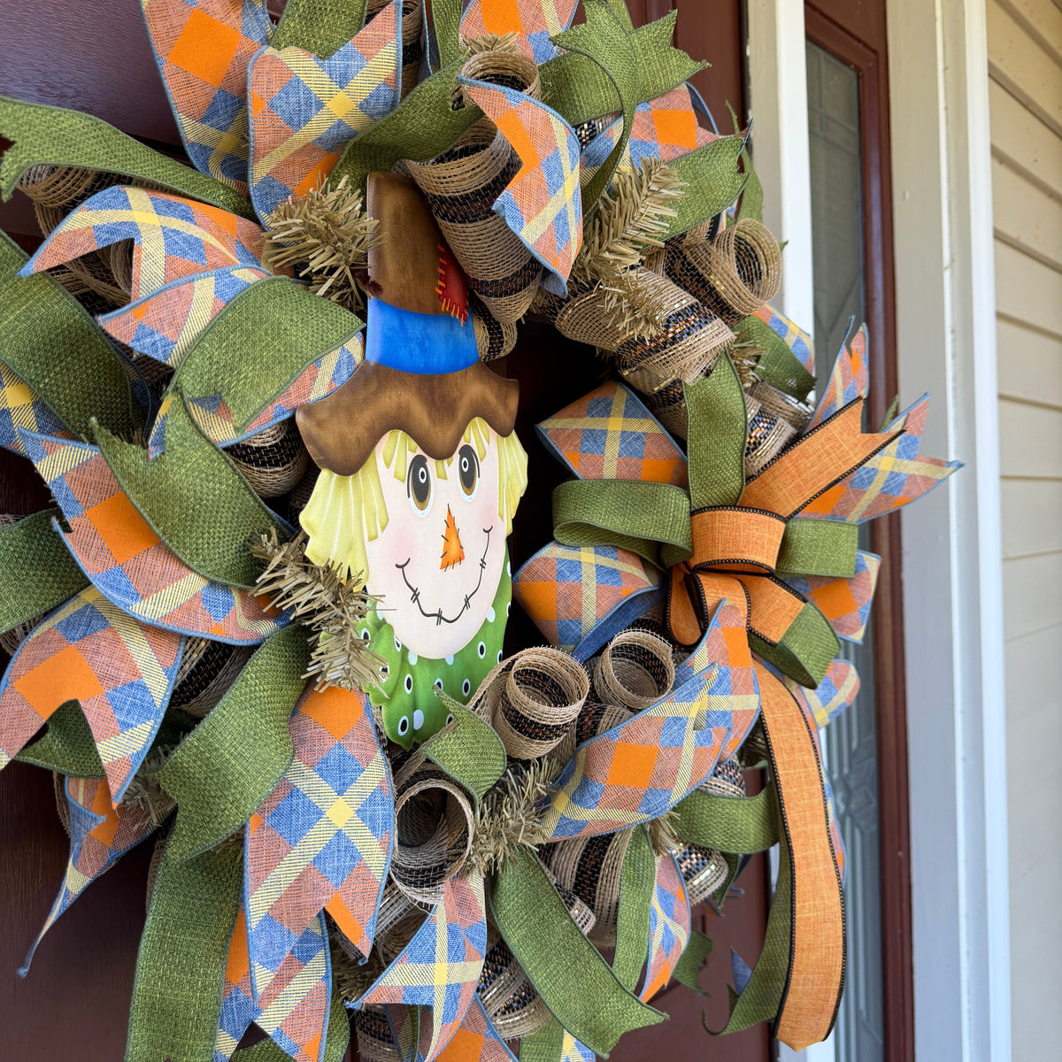 colorful scarecrow fall wreath. With its smiling scarecrow, warm autumn tones, and playful plaid ribbon, this wreath feels lighthearted, friendly, and full of seasonal charm.