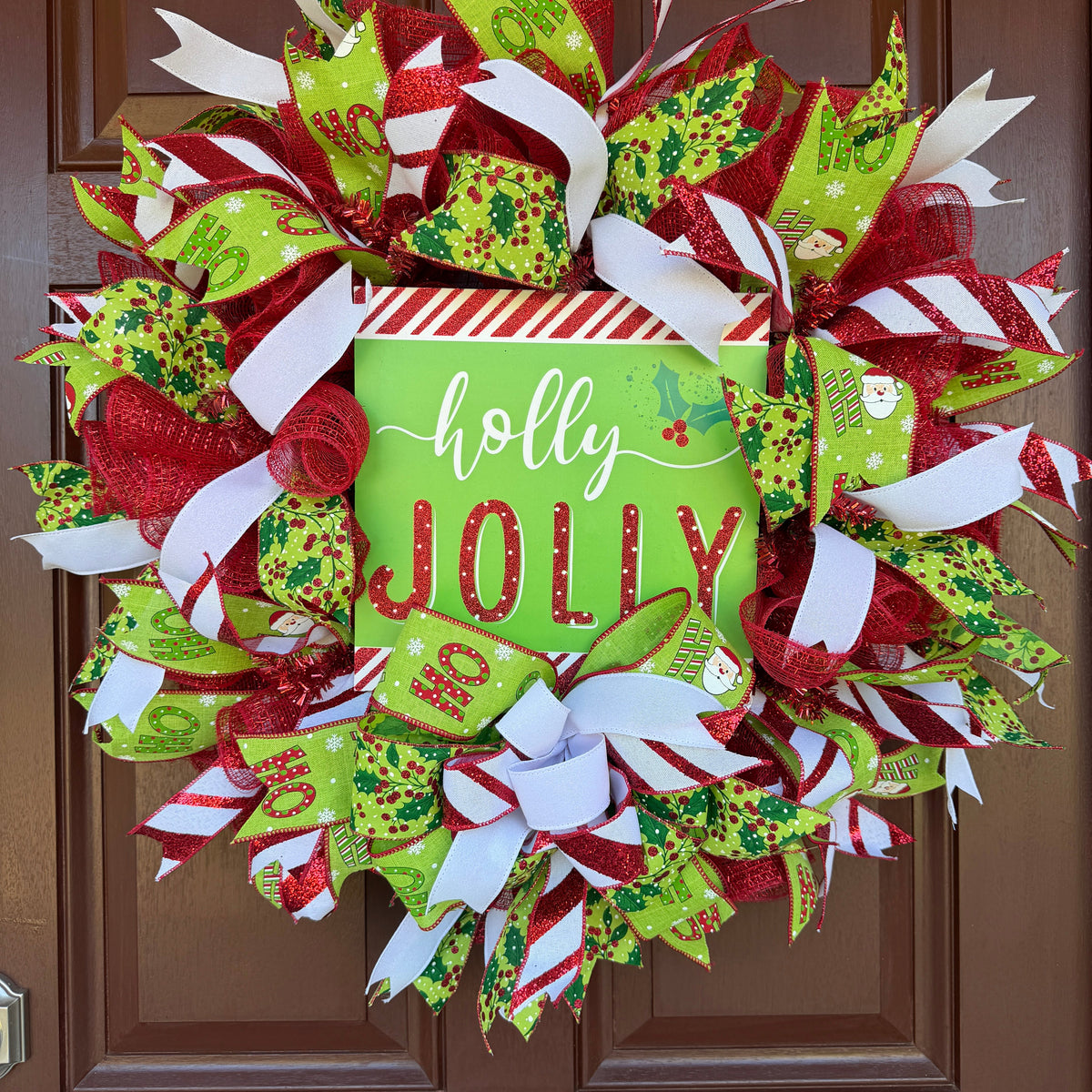 A festive Christmas wreath made of red, green, and white mesh ribbons with candy cane, holly, and snowflake patterns, featuring a central square sign that reads 'holly JOLLY' in white and red lettering.