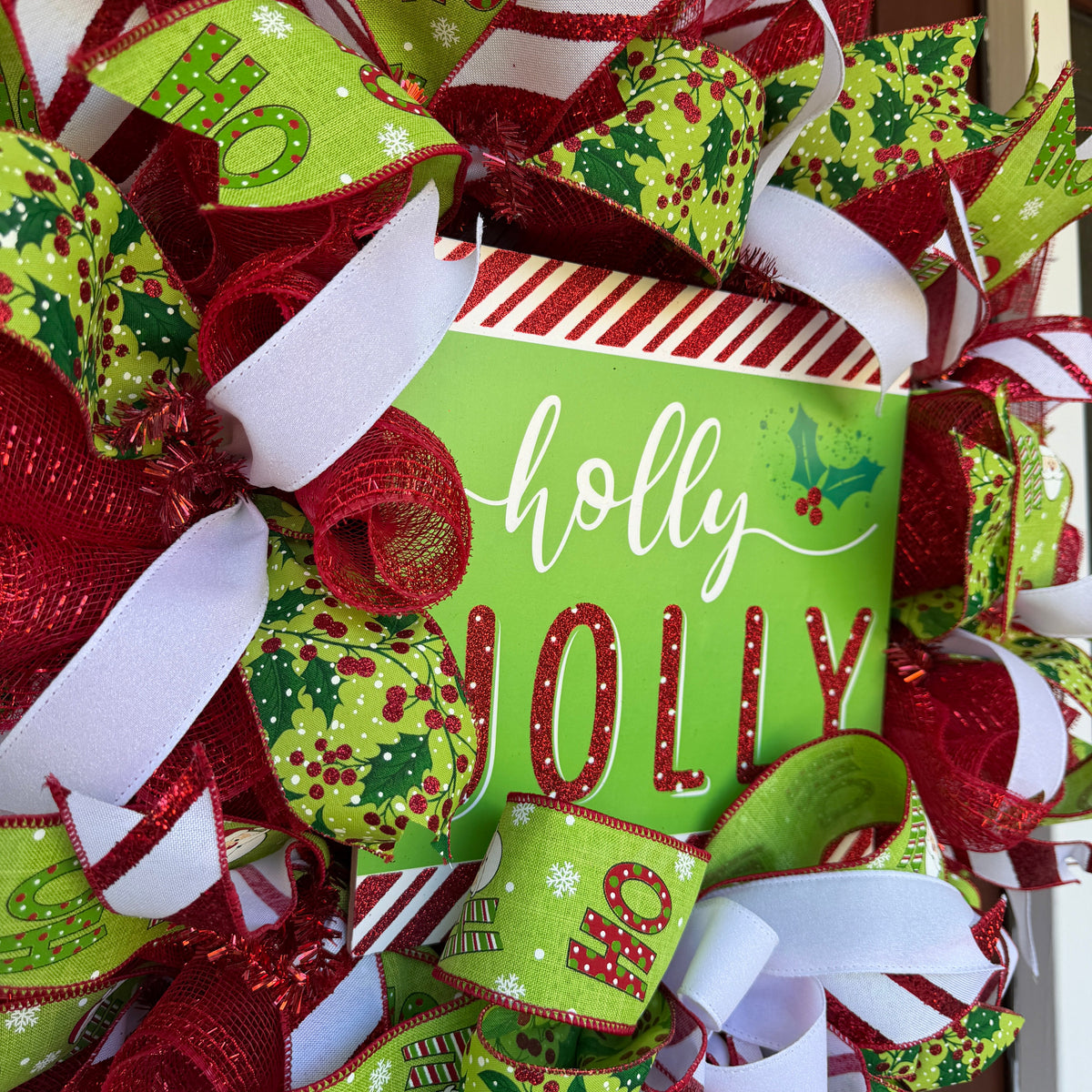 A festive Christmas wreath made of red, green, and white mesh ribbons with candy cane, holly, and snowflake patterns, featuring a central square sign that reads 'holly JOLLY' in white and red lettering.