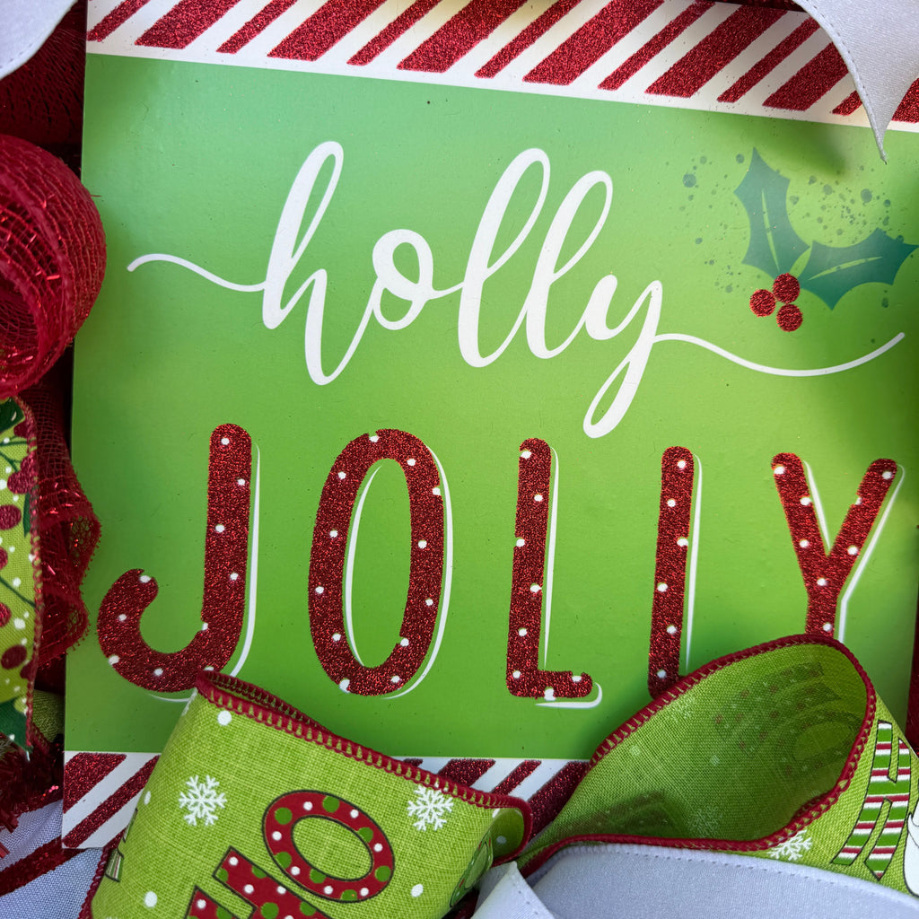 A festive Christmas wreath made of red, green, and white mesh ribbons with candy cane, holly, and snowflake patterns, featuring a central square sign that reads 'holly JOLLY' in white and red lettering.
