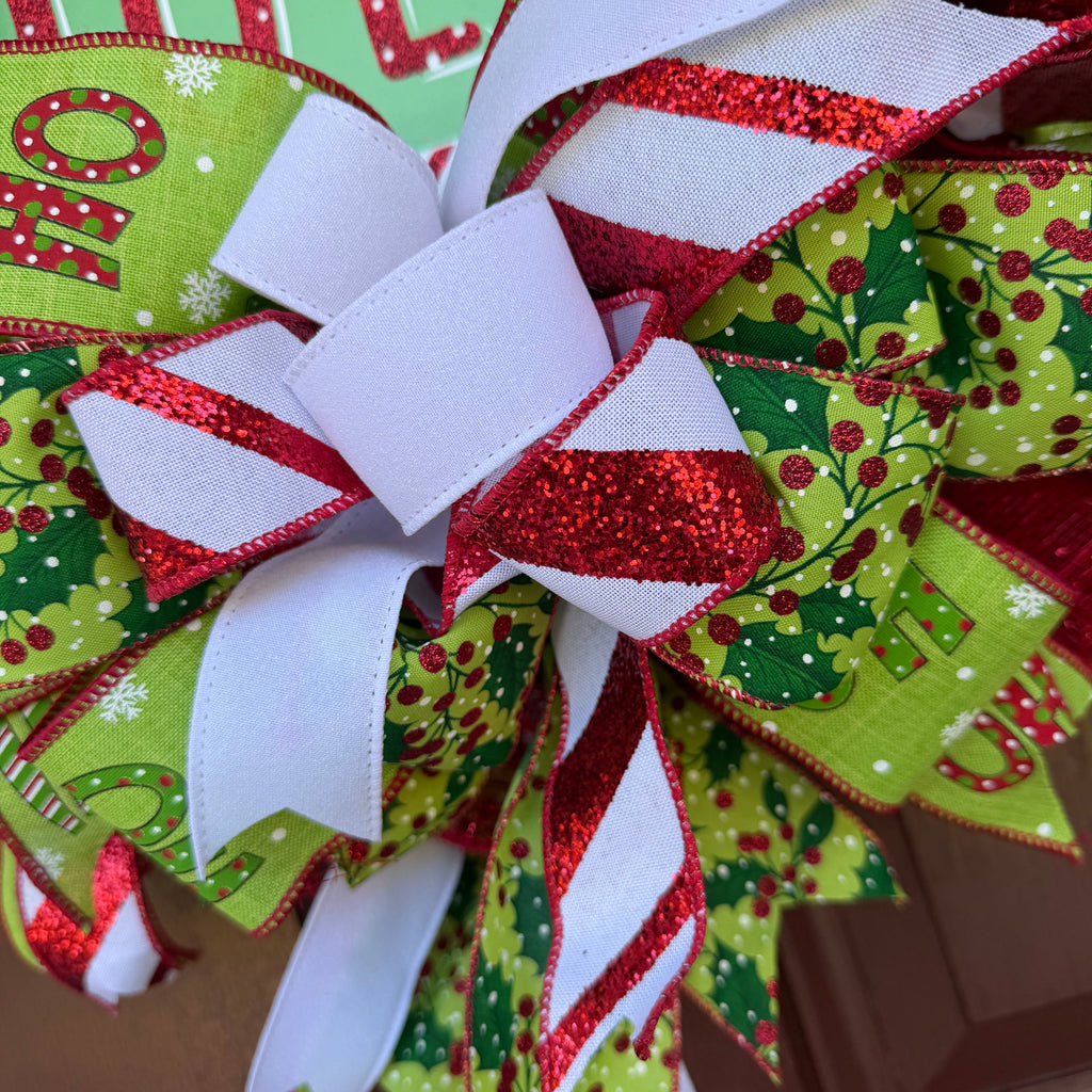 A festive Christmas wreath made of red, green, and white mesh ribbons with candy cane, holly, and snowflake patterns, featuring a central square sign that reads 'holly JOLLY' in white and red lettering.