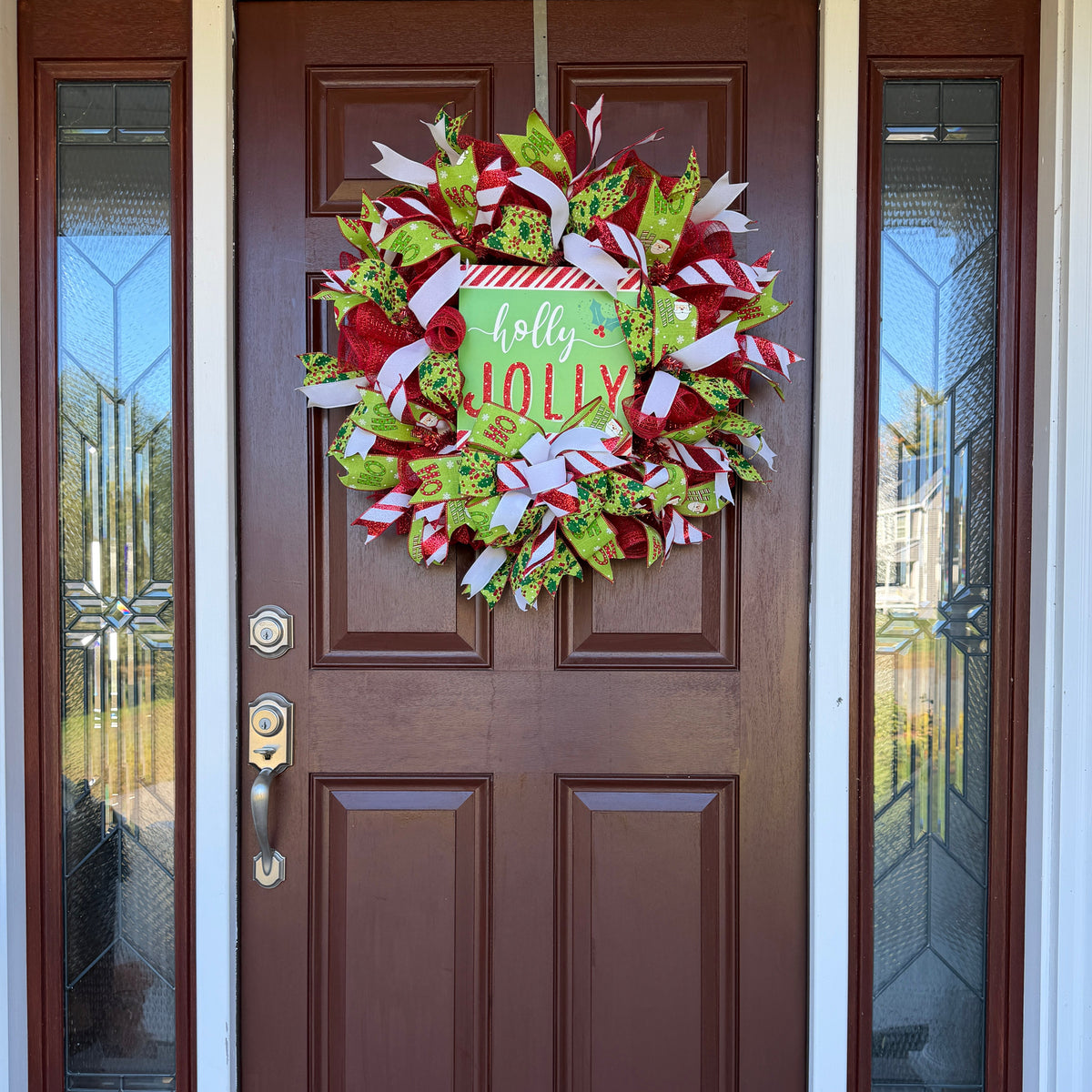 A festive Christmas wreath made of red, green, and white mesh ribbons with candy cane, holly, and snowflake patterns, featuring a central square sign that reads 'holly JOLLY' in white and red lettering.