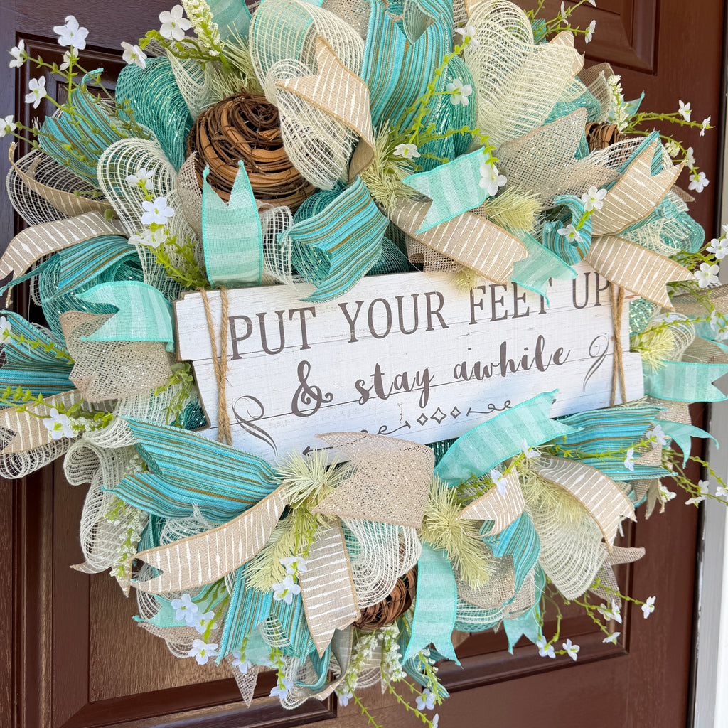 Handmade coastal welcome wreath with aqua blue, cream, and beige ribbons, floral accents, and a farmhouse-style welcome sign for beach house or summer front door decor.
