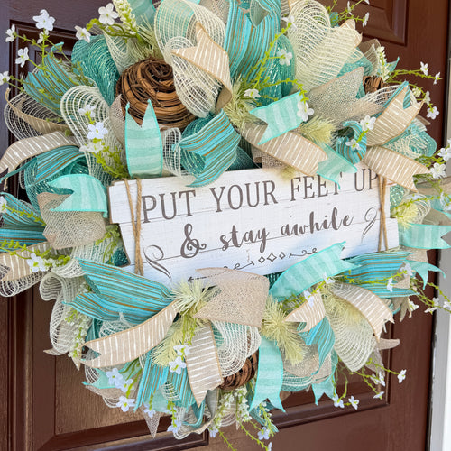 Handmade coastal welcome wreath with aqua blue, cream, and beige ribbons, floral accents, and a farmhouse-style welcome sign for beach house or summer front door decor.
