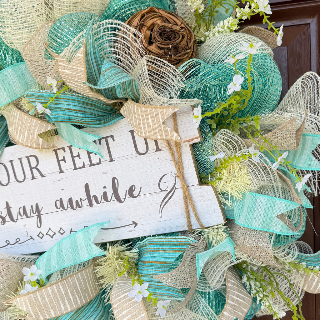 Handmade coastal welcome wreath with aqua blue, cream, and beige ribbons, floral accents, and a farmhouse-style welcome sign for beach house or summer front door decor.