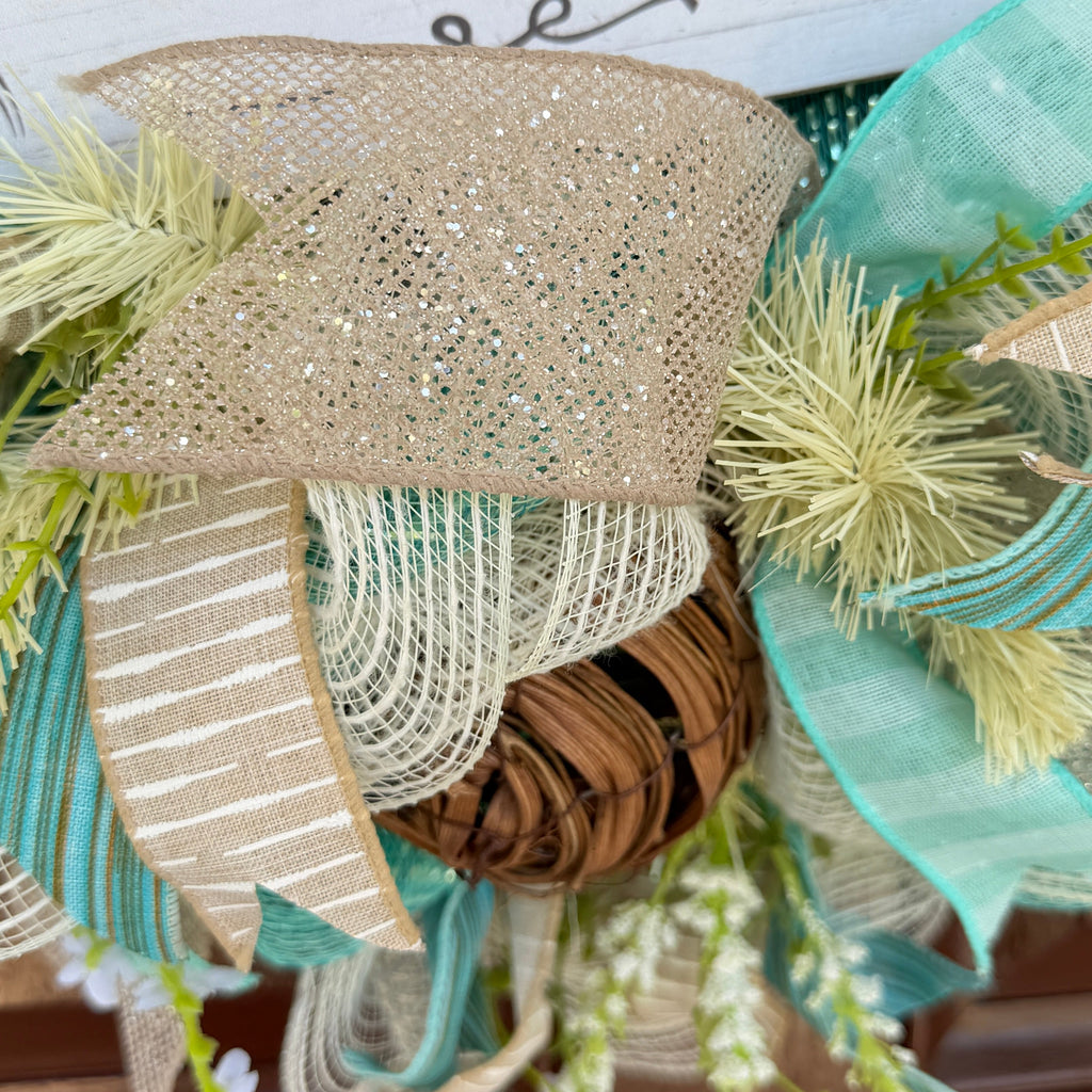 Handmade coastal welcome wreath with aqua blue, cream, and beige ribbons, floral accents, and a farmhouse-style welcome sign for beach house or summer front door decor.