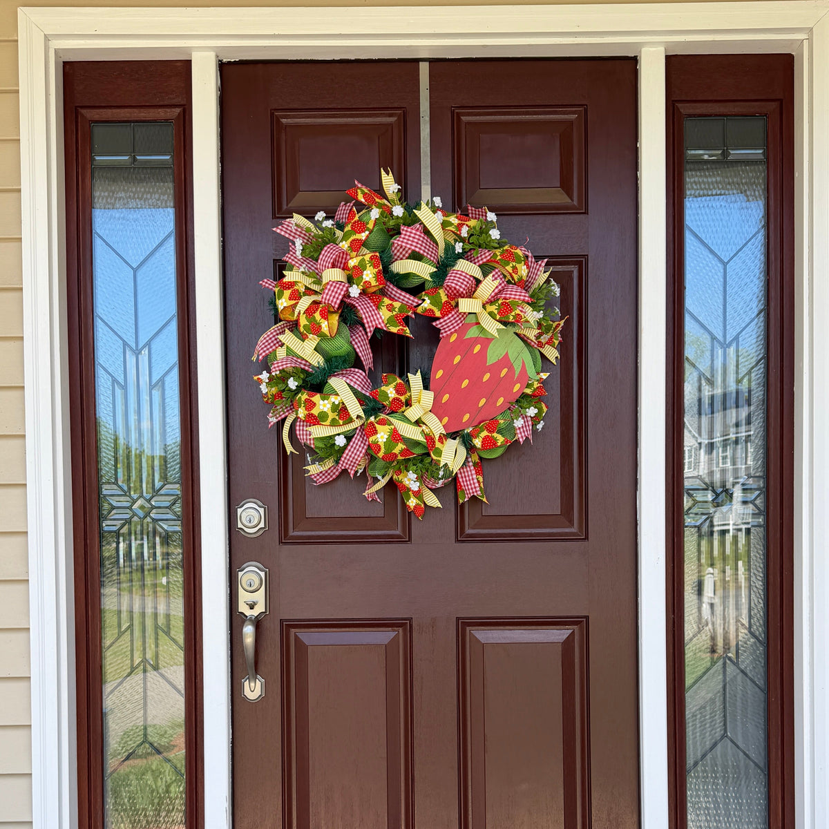 Handmade strawberry summer wreath featuring red gingham ribbons, strawberry print accents, greenery, and a large strawberry centerpiece hanging on a front door.