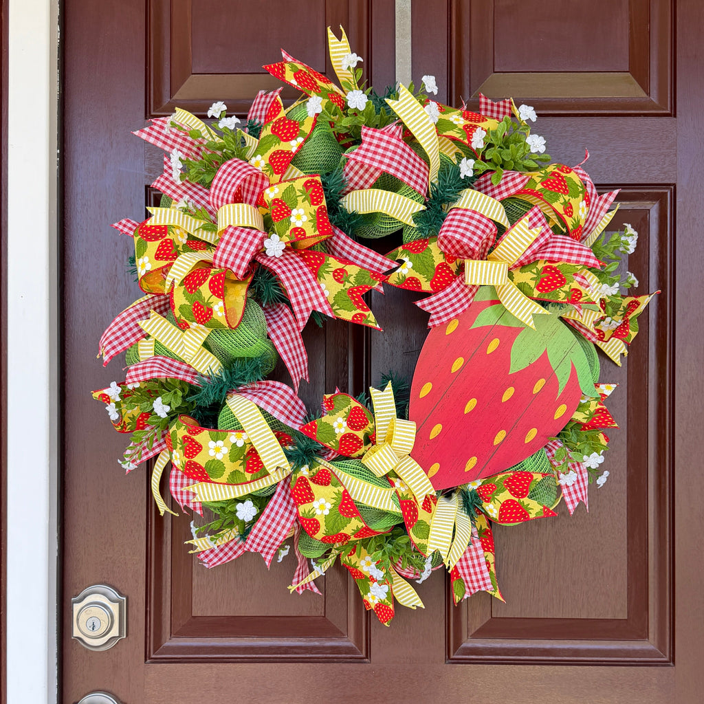 Handmade strawberry summer wreath featuring red gingham ribbons, strawberry print accents, greenery, and a large strawberry centerpiece hanging on a front door.