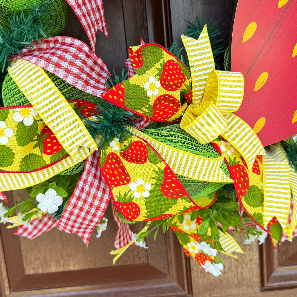 Handmade strawberry summer wreath featuring red gingham ribbons, strawberry print accents, greenery, and a large strawberry centerpiece hanging on a front door.