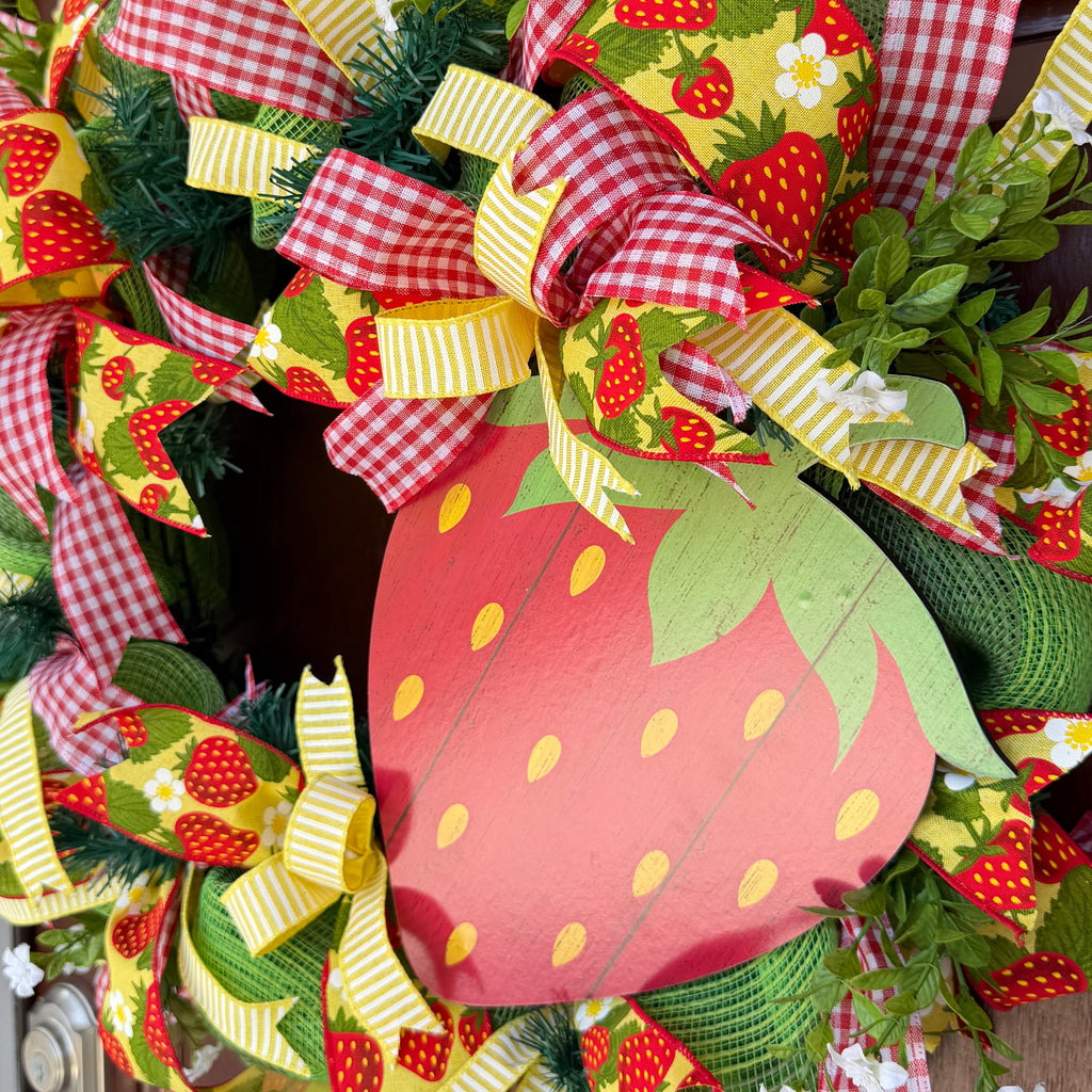 Handmade strawberry summer wreath featuring red gingham ribbons, strawberry print accents, greenery, and a large strawberry centerpiece hanging on a front door.