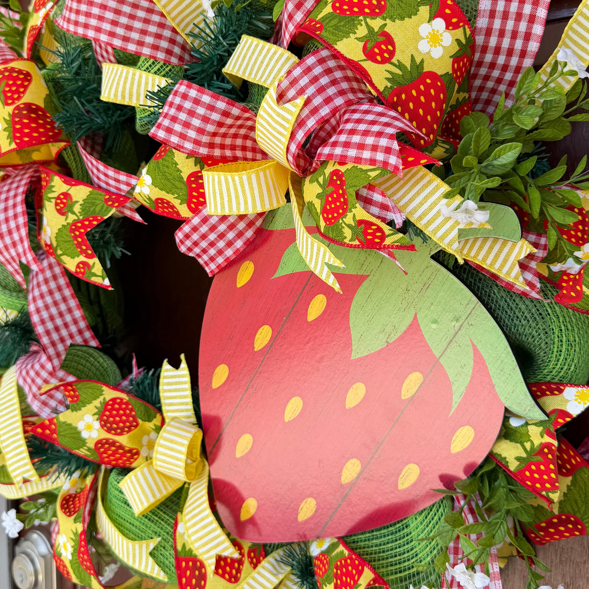 Handmade strawberry summer wreath featuring red gingham ribbons, strawberry print accents, greenery, and a large strawberry centerpiece hanging on a front door.