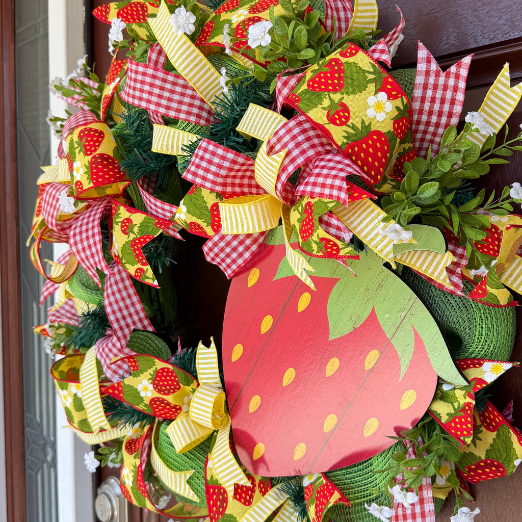 Handmade strawberry summer wreath featuring red gingham ribbons, strawberry print accents, greenery, and a large strawberry centerpiece hanging on a front door.