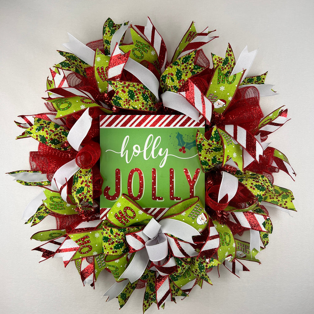A festive Christmas wreath made of red, green, and white mesh ribbons with candy cane, holly, and snowflake patterns, featuring a central square sign that reads 'holly JOLLY' in white and red lettering.