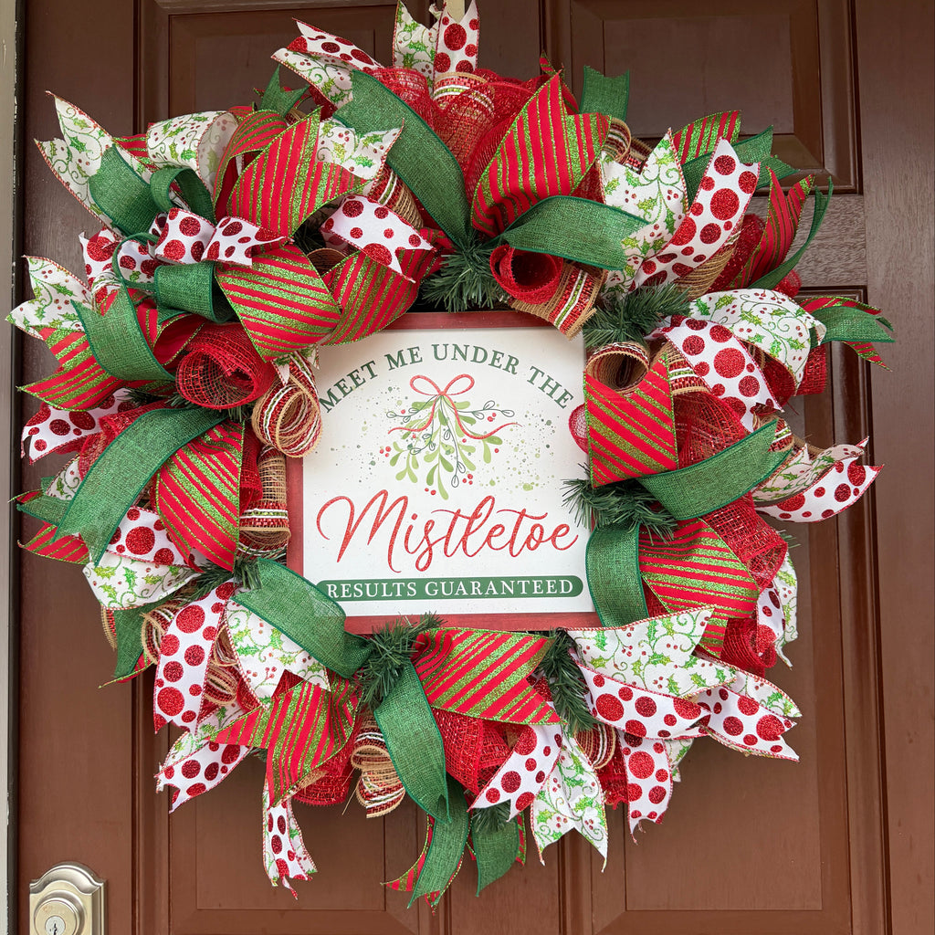 Handmade red and green Christmas deco mesh wreath with glittery ribbons, holly accents, and Meet Me Under the Mistletoe sign