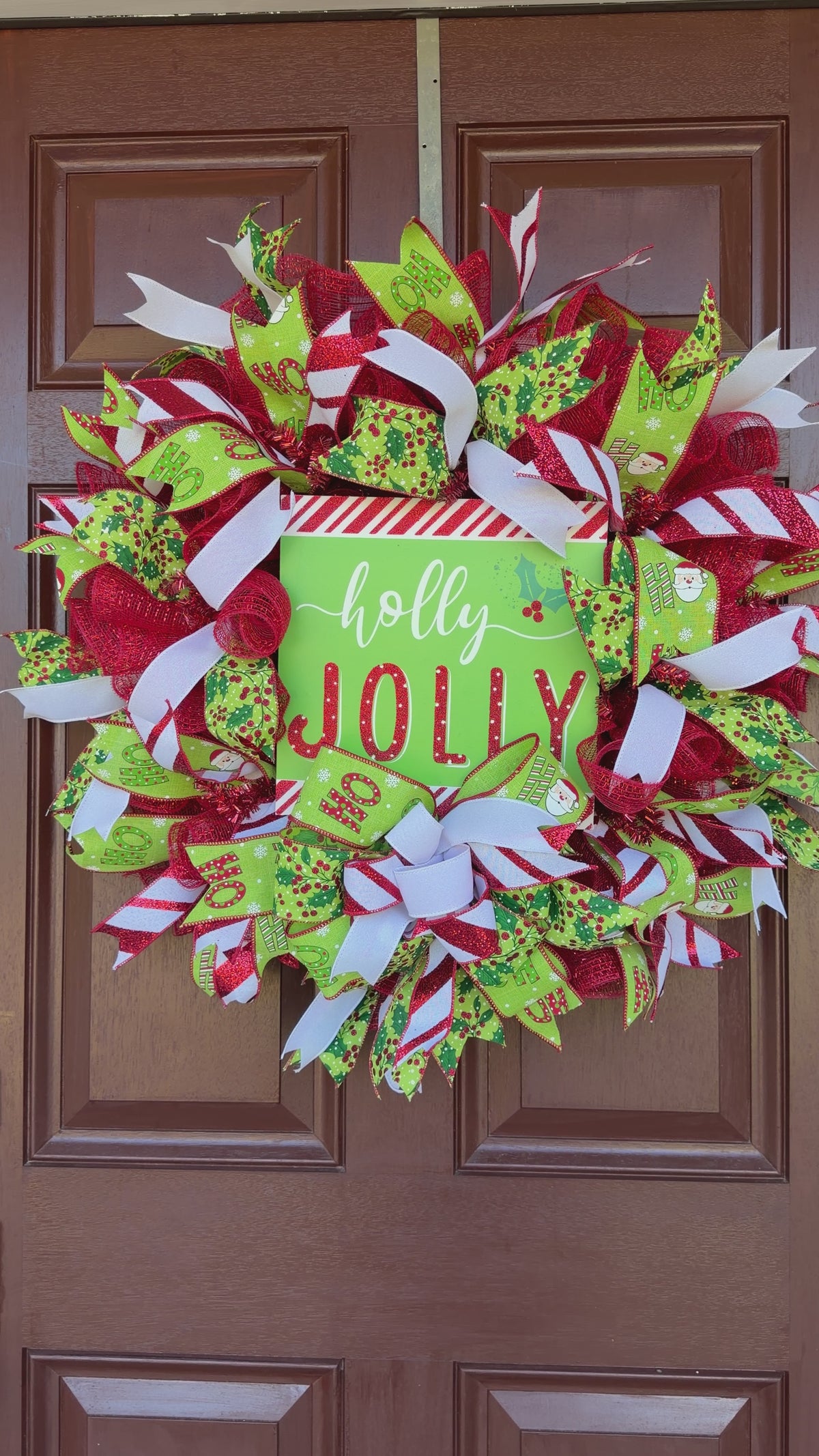 A festive Christmas wreath made of red, green, and white mesh ribbons with candy cane, holly, and snowflake patterns, featuring a central square sign that reads 'holly JOLLY' in white and red lettering.