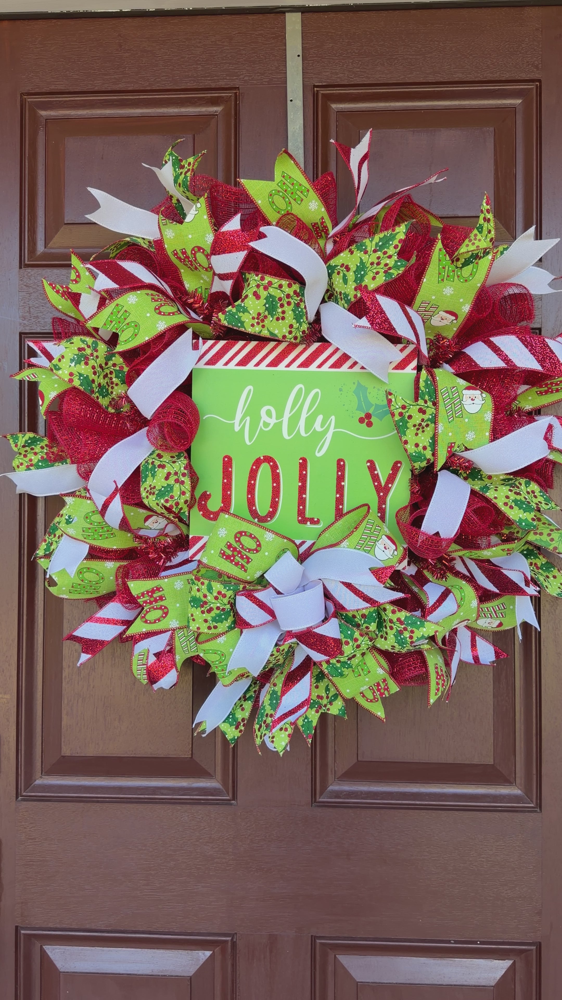 A festive Christmas wreath made of red, green, and white mesh ribbons with candy cane, holly, and snowflake patterns, featuring a central square sign that reads 'holly JOLLY' in white and red lettering.