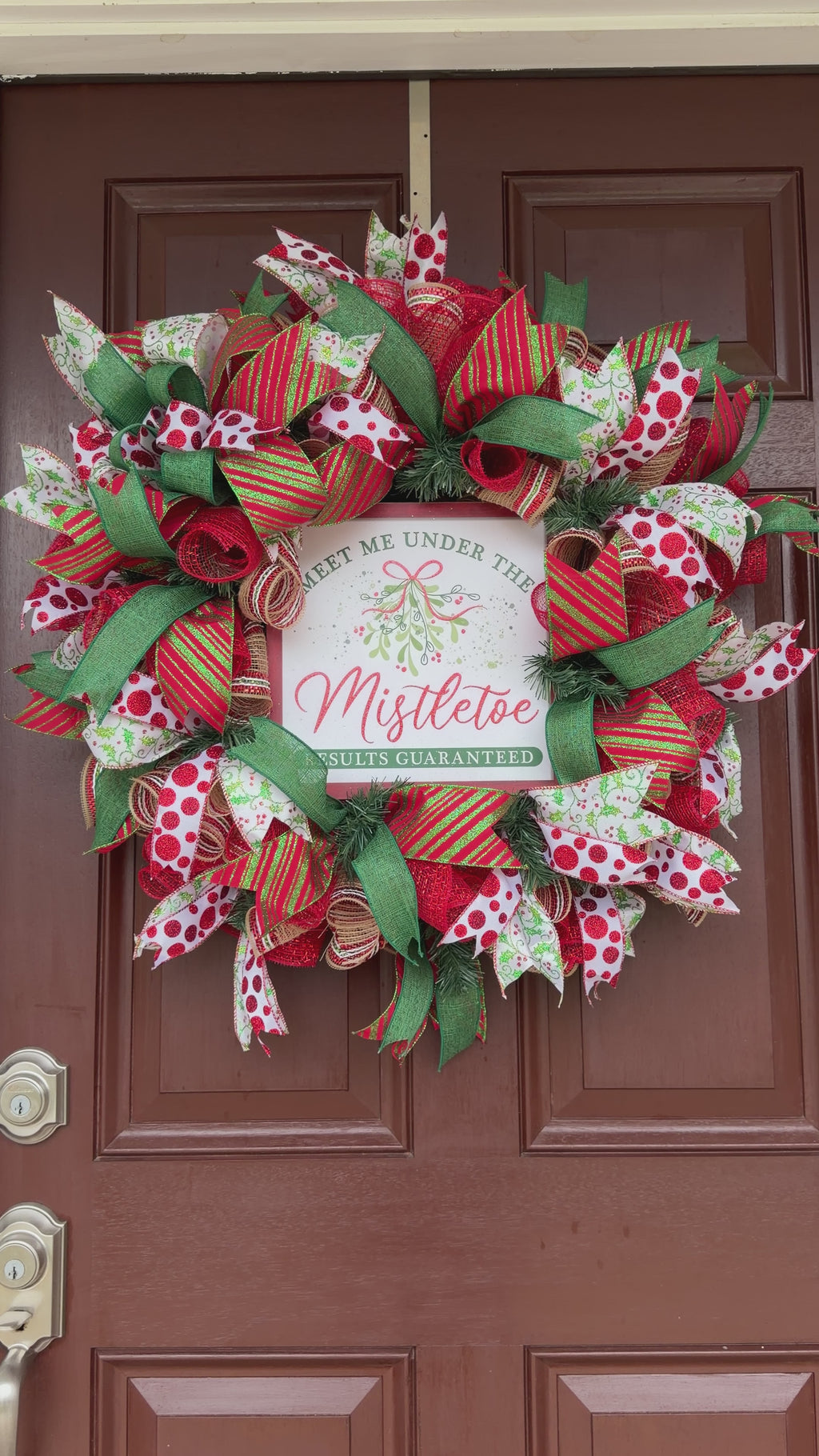 Handmade red and green Christmas deco mesh wreath with glittery ribbons, holly accents, and Meet Me Under the Mistletoe sign