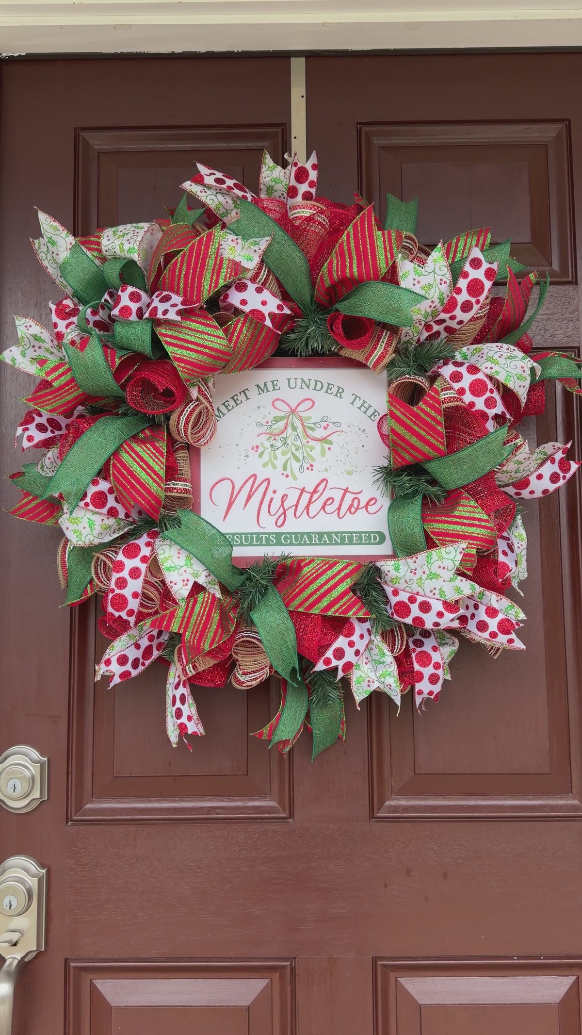 Handmade red and green Christmas deco mesh wreath with glittery ribbons, holly accents, and Meet Me Under the Mistletoe sign