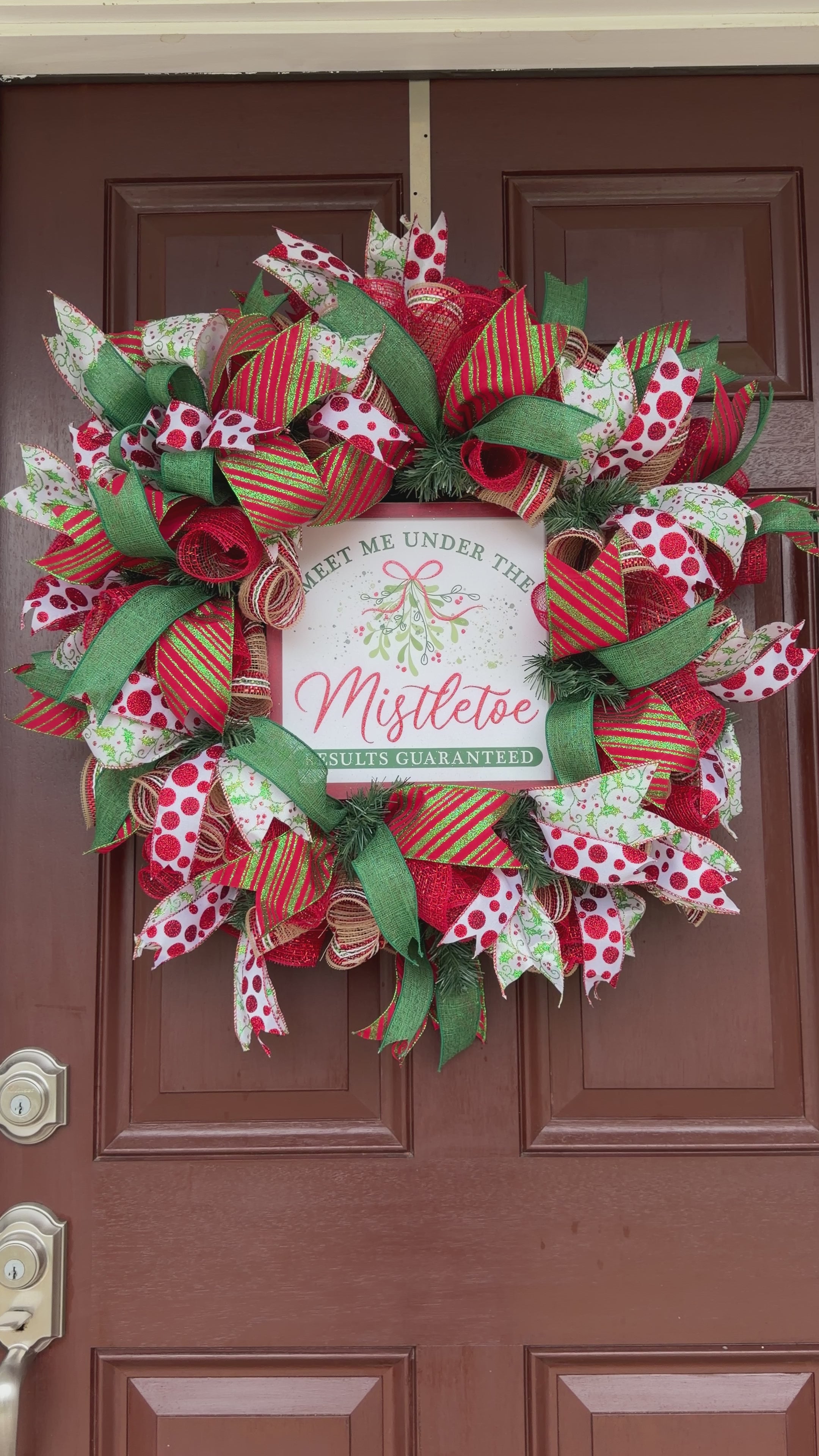 Handmade red and green Christmas deco mesh wreath with glittery ribbons, holly accents, and Meet Me Under the Mistletoe sign