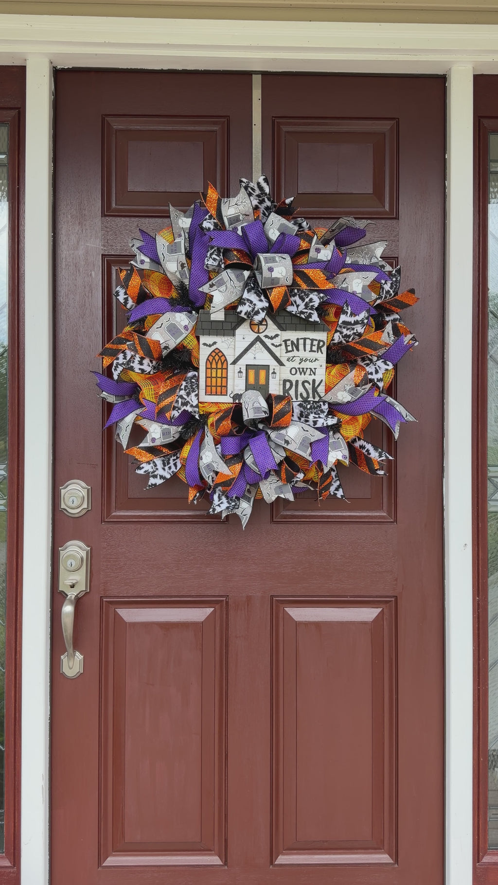 A Halloween-themed mesh wreath hanging on a brown front door. The wreath features purple, orange, black, and white ribbons with a central white house sign that reads 'ENTER at your OWN RISK' with a pumpkin and bats detail.