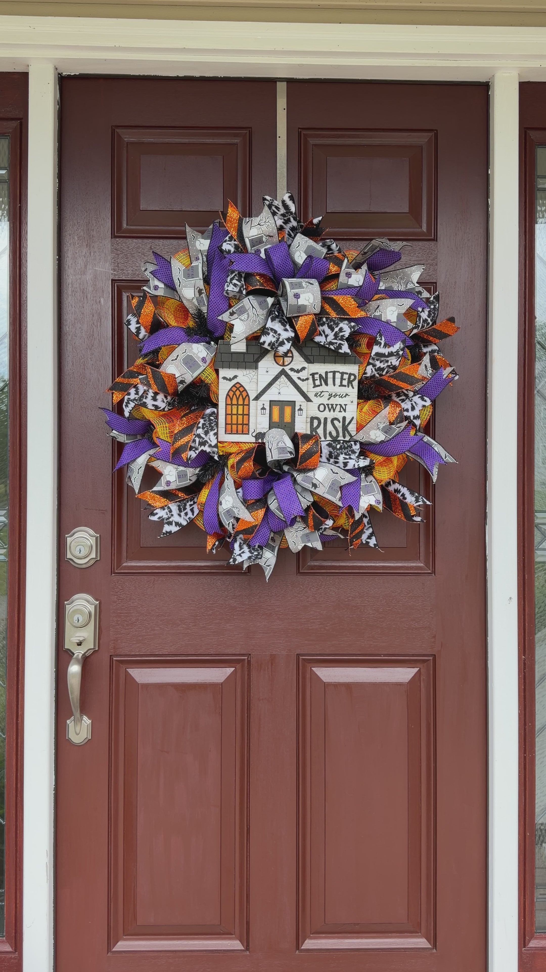 A Halloween-themed mesh wreath hanging on a brown front door. The wreath features purple, orange, black, and white ribbons with a central white house sign that reads 'ENTER at your OWN RISK' with a pumpkin and bats detail.
