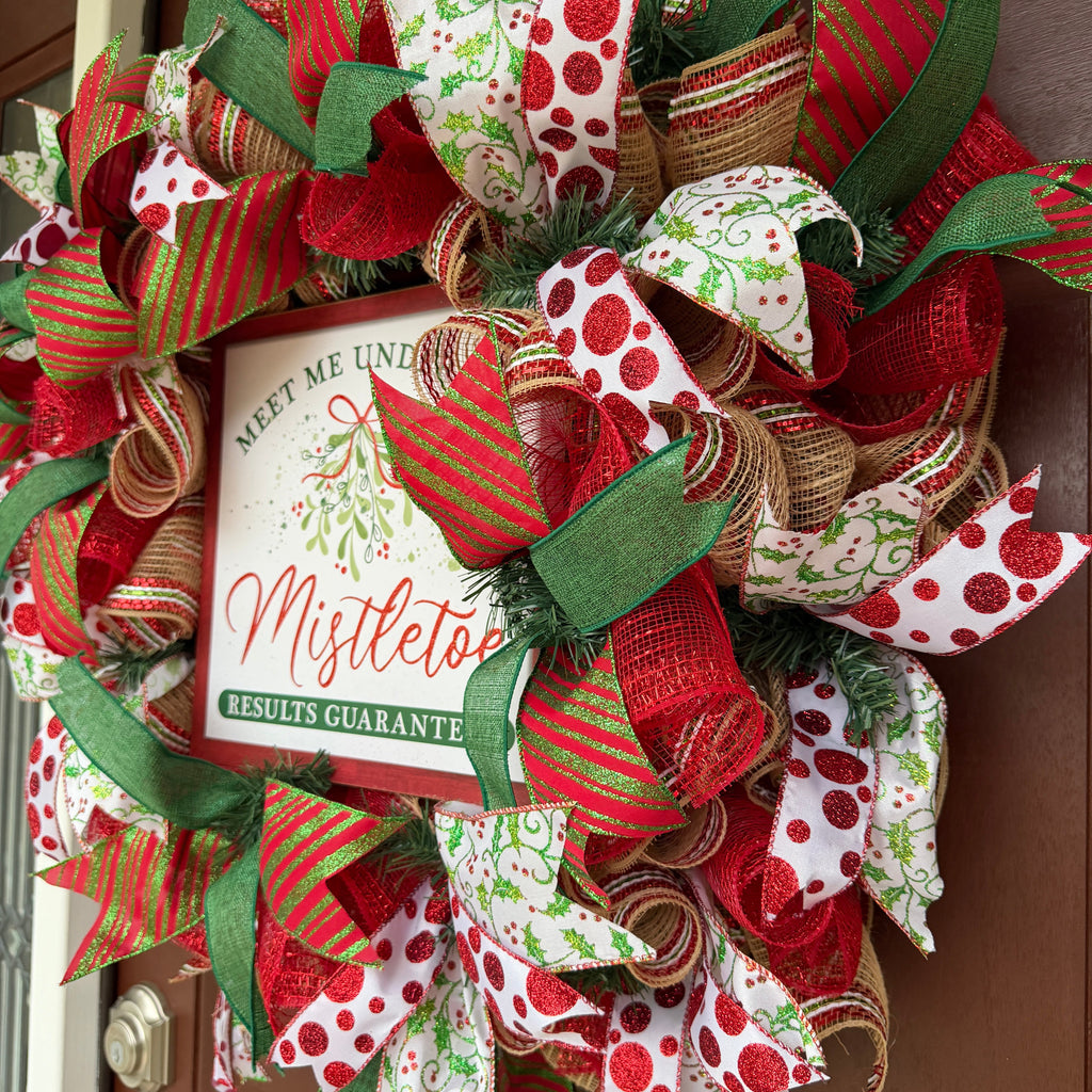 Handmade red and green Christmas deco mesh wreath with glittery ribbons, holly accents, and Meet Me Under the Mistletoe sign