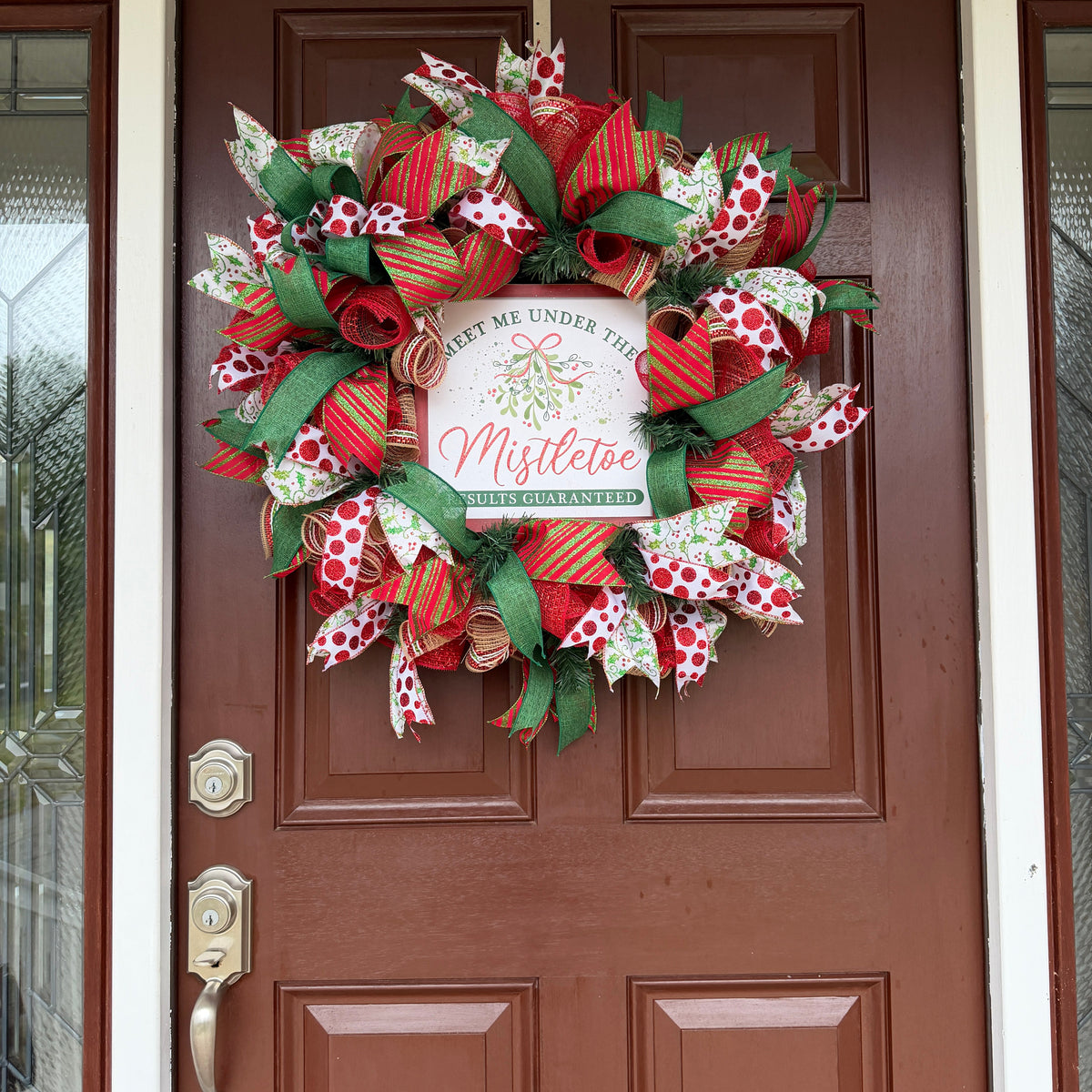 Handmade red and green Christmas deco mesh wreath with glittery ribbons, holly accents, and Meet Me Under the Mistletoe sign